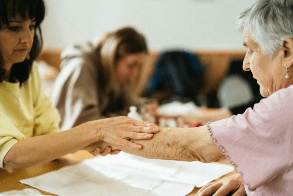 A healthcare worker gently holds the arm of an elderly woman during a care appointment.