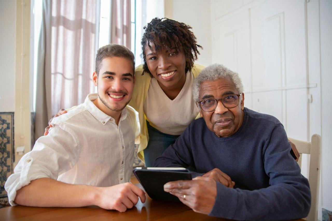 A senior man reviews information on a tablet with two younger family members at a dining table.